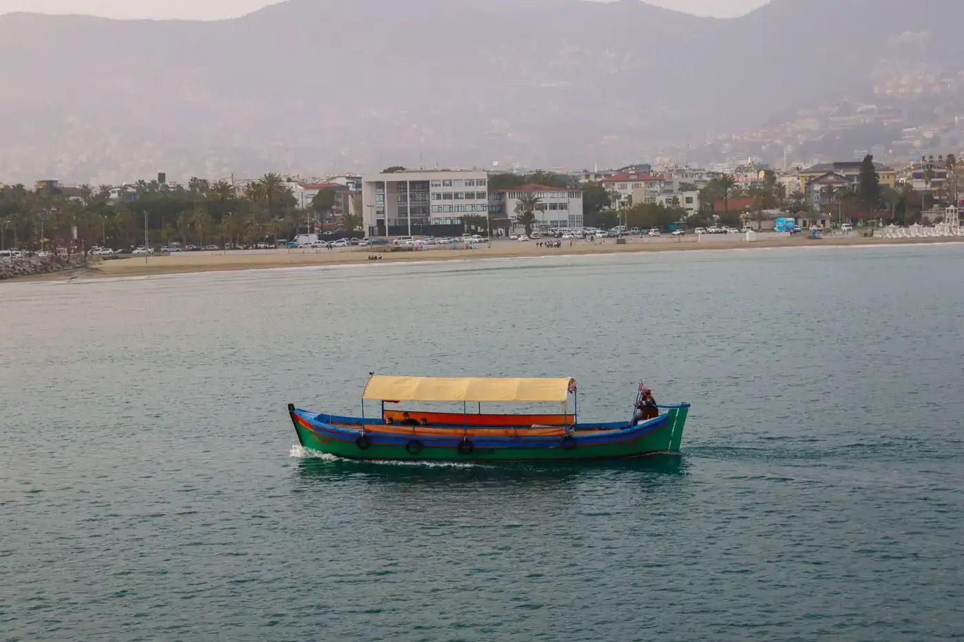 A colorful traditional boat sails on Antalya's coastal waters, with the cityscape in the background.