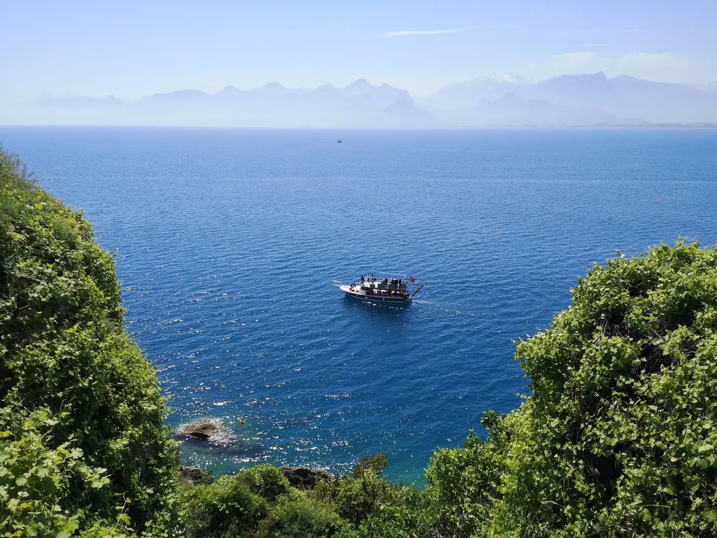 A picturesque boat sailing in the blue waters of Antalya, Turkey, with lush greenery and mountains.