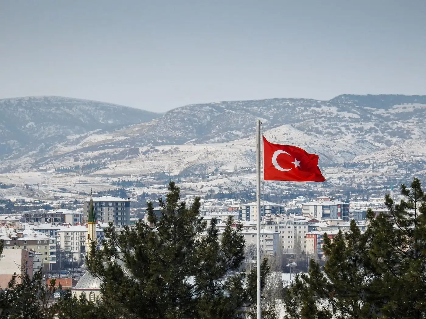 Scenic view of Turkish flag waving over snowy cityscape of Mengen, Bolu, with mountains in the background.
