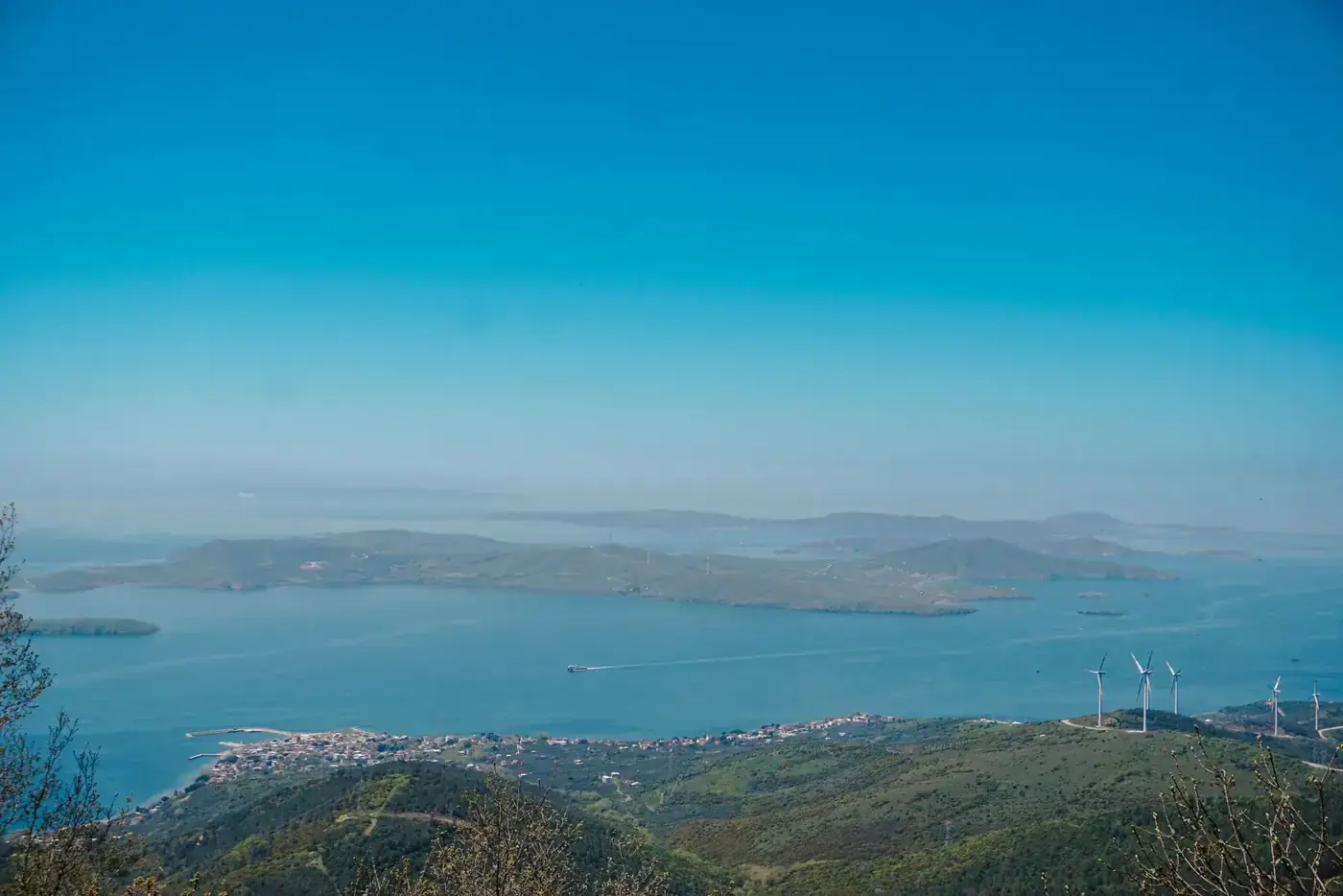 Aerial view of the tranquil sea and islands in Balıkesir, Türkiye, with wind turbines on a hill under a clear blue sky.
