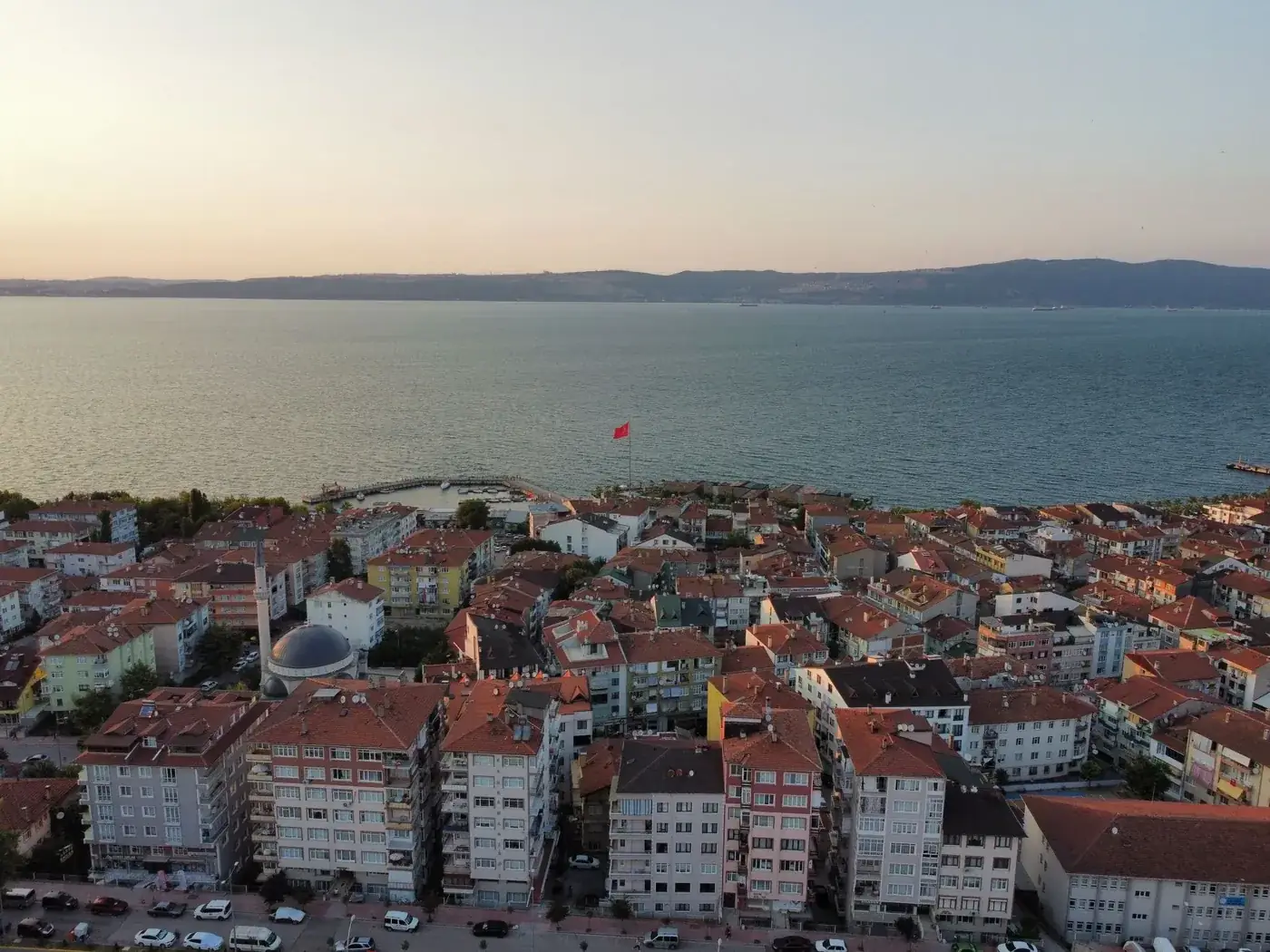 Aerial view of a coastal city with red rooftops at sunset overlooking the ocean and distant hills.