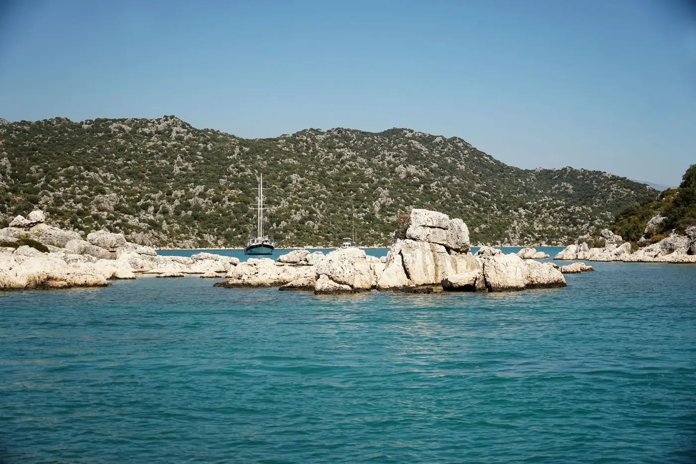 Calm sea with rocky coastlines and a sailboat against hilly terrain.
