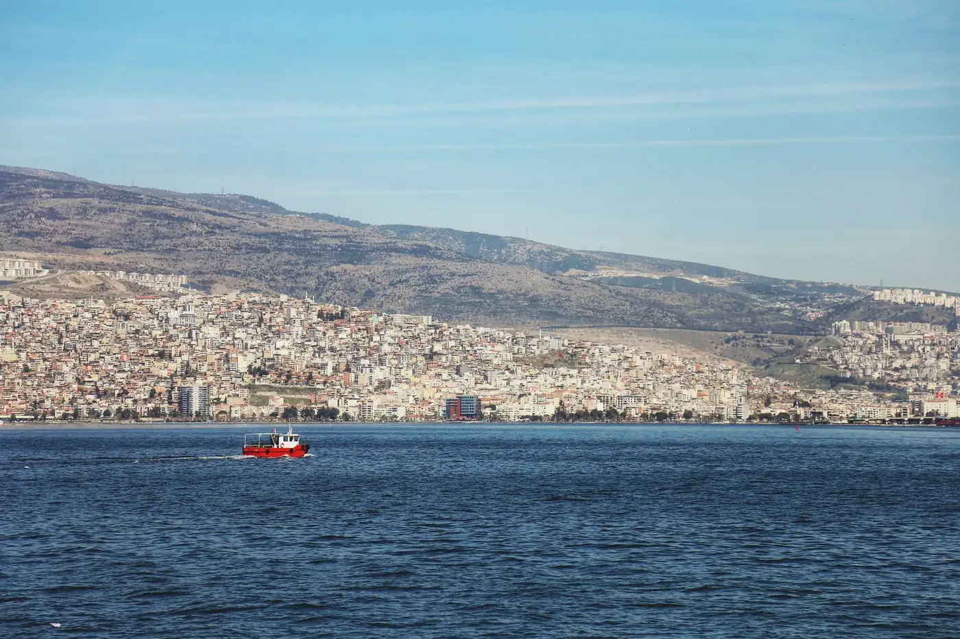 Red boat sailing in Izmir Bay with a sprawling cityscape and mountains in the background.